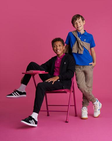 two kids on a fushia background, one is sitting and the other one is standing both smiling
